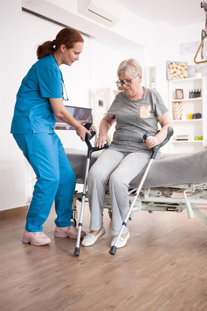 Enfermera ayudando a una anciana a sentarse en la cama en un hogar de ancianos después de caminar con muletas.