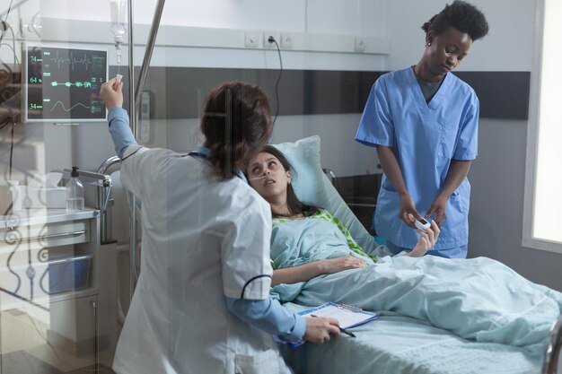 Enfermera colocando oxímetro de pulso en el dedo del paciente mientras el médico configura el catéter de la vía central con medicación por goteo intravenoso. Mujer en cama de hospital con baja saturación de oxígeno recibiendo atención médica.