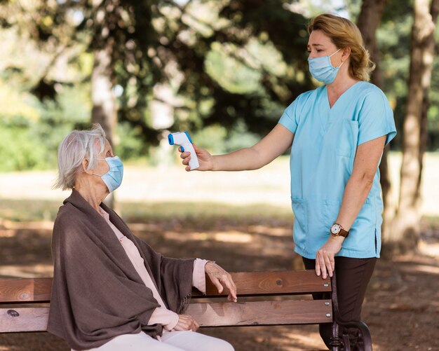 Enfermera comprobando la temperatura de la mujer mayor al aire libre en un banco