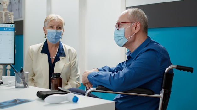 Enfermera consultando a un anciano con discapacidad física en la cita de control, hablando con el paciente y la esposa con máscaras faciales. Asistente de reunión con usuario de silla de ruedas, dando apoyo a la discapacidad.