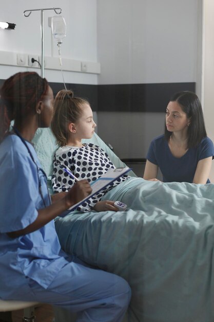 Enfermera consultando a una hija pequeña hospitalizada enferma descansando en la cama del paciente de la sala de la clínica de atención médica pediátrica. Niño enfermo recuperándose de una enfermedad mientras el personal médico realiza un chequeo del estado de salud.