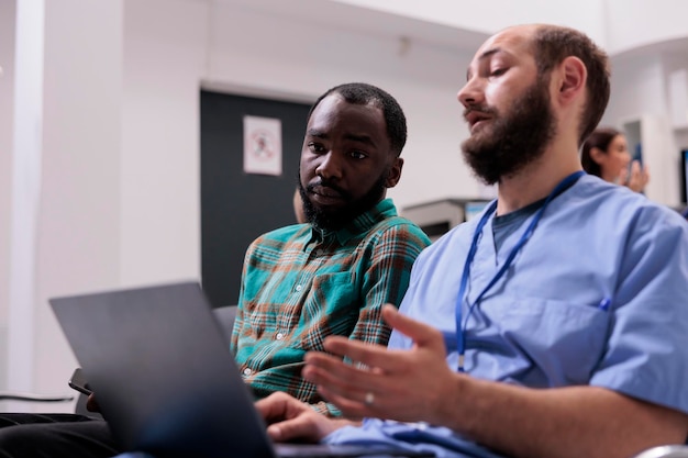 Enfermera consultando a un paciente afroamericano en el área de espera, usando una computadora portátil para explicar el diagnóstico de la enfermedad y el tratamiento de recuperación en el vestíbulo de recepción. Gente hablando de atención médica en las instalaciones.