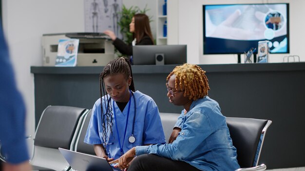 Enfermera explicando la diangosis en una laptop al paciente en el área de espera del centro médico. Especialista en atención médica que usa computadora en el examen de consulta con un adulto joven en los asientos de la sala de espera.