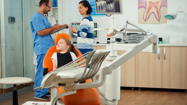 Foto gratuita enfermera invitando a paciente niño en consulta dental, pediatra hablando con niña. asistente de hombre hablando con la madre preparándose para el examen estomatológico en la oficina del ortodoncista