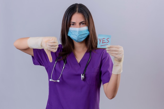 Enfermera joven disgustada con guantes de máscara protectora uniforme médico y con estetoscopio sosteniendo papel recordatorio con la palabra sí mostrando el pulgar hacia abajo