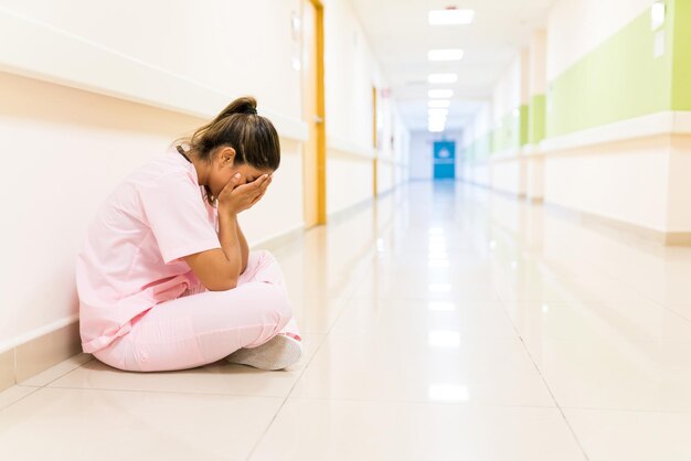 Enfermera joven estresada que cubre la cara mientras está sentada en el suelo en el pasillo del hospital