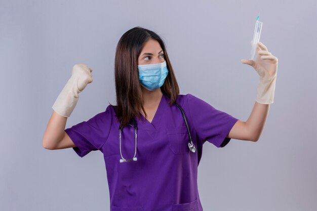 Enfermera joven con guantes de máscara protectora uniforme médico y con estetoscopio sosteniendo una jeringa levantando el puño como un ganador mostrando fuerza