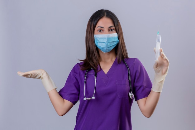 Enfermera joven con guantes de máscara protectora uniforme médico y con estetoscopio sosteniendo una jeringa mirando desorientado y confundido de pie con el brazo levantado