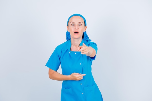 Enfermera joven sorprendida está apuntando a la cámara con el dedo índice sobre fondo blanco.