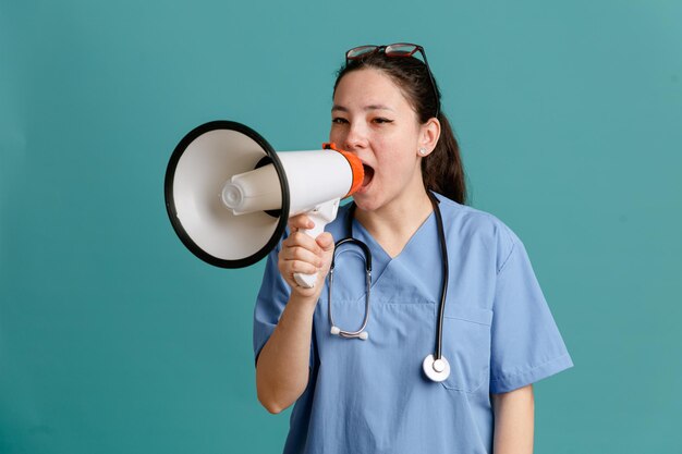 Enfermera joven en uniforme médico con estetoscopio alrededor del cuello gritando en megáfono mirando confiada de pie sobre fondo azul.