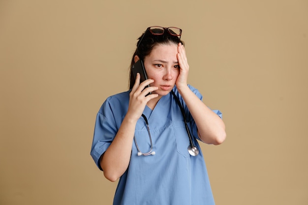 Enfermera joven en uniforme médico con estetoscopio alrededor del cuello hablando por teléfono móvil mirando confundida sosteniendo la mano en su cabeza por error de pie sobre fondo marrón