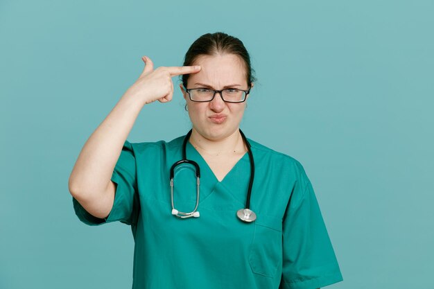 Enfermera joven en uniforme médico con estetoscopio alrededor del cuello mirando a la cámara confundida y decepcionada señalando con el dedo índice a su sien de pie sobre fondo azul