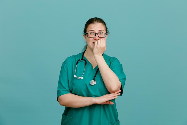 Enfermera joven en uniforme médico con estetoscopio alrededor del cuello mirando a la cámara estresada y nerviosa mordiéndose las uñas de pie sobre fondo azul.