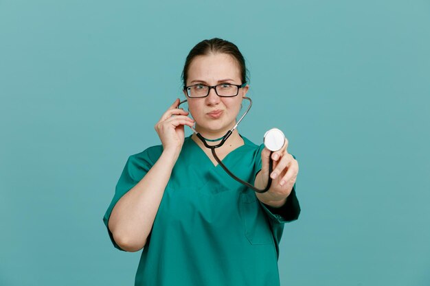 Enfermera joven en uniforme médico con estetoscopio alrededor del cuello mirando a la cámara con expresión segura de pie sobre fondo azul.