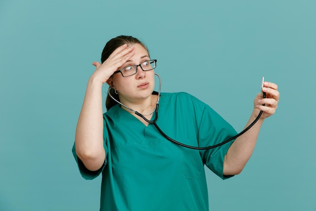 Enfermera joven en uniforme médico con estetoscopio alrededor del cuello que parece confundida y sorprendida de pie sobre fondo azul.