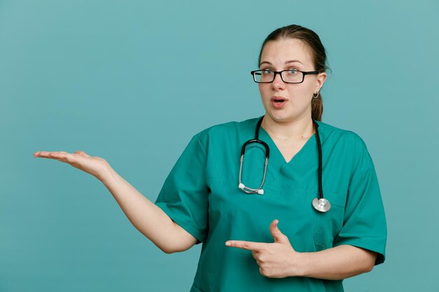 Enfermera joven en uniforme médico con estetoscopio alrededor del cuello que presenta algo con la mano en la mano apuntando con el dedo índice hacia un lado mirando confundida de pie sobre fondo azul