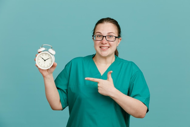 Enfermera joven con uniforme médico con estetoscopio alrededor del cuello sosteniendo un despertador apuntándolo con el dedo índice feliz y positiva sonriendo alegremente de pie sobre fondo azul