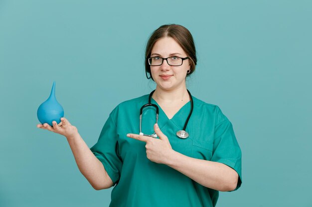 Enfermera joven en uniforme médico con estetoscopio alrededor del cuello sosteniendo enema mirando a la cámara sonriendo confiada apuntando con el dedo índice al enema de pie sobre fondo azul