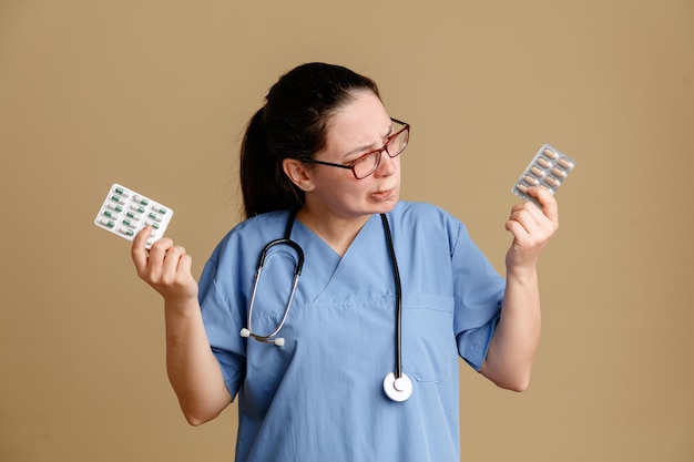 Enfermera joven en uniforme médico con estetoscopio alrededor del cuello sosteniendo pastillas que parecen confundidas tratando de elegir de pie sobre fondo marrón