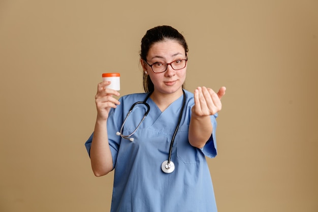 Enfermera joven en uniforme médico con estetoscopio alrededor del cuello sosteniendo un pequeño frasco de prueba mirando a la cámara sonriendo confiada haciendo gesto de venir aquí con el brazo de pie sobre fondo marrón