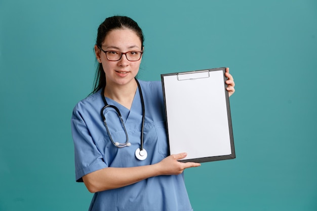 Enfermera joven en uniforme médico con estetoscopio alrededor del cuello sosteniendo portapapeles con página en blanco mirando a la cámara sonriendo confiada de pie sobre fondo azul