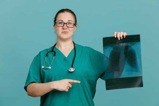 Enfermera joven con uniforme médico con estetoscopio alrededor del cuello sosteniendo una radiografía pulmonar apuntando con el dedo índice mirando confundida y disgustada de pie sobre fondo azul