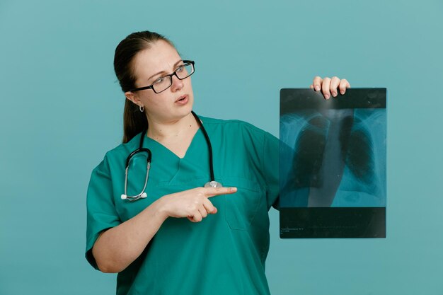 Enfermera joven en uniforme médico con estetoscopio alrededor del cuello sosteniendo radiografía pulmonar apuntando con el dedo índice mirando preocupada de pie sobre fondo azul