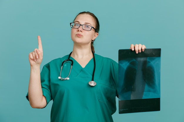 Foto gratuita enfermera joven con uniforme médico con estetoscopio alrededor del cuello sosteniendo una radiografía pulmonar que parece confiada mostrando el dedo índice con buenas noticias de pie sobre fondo azul