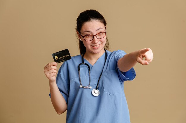 Enfermera joven en uniforme médico con estetoscopio alrededor del cuello con tarjeta de crédito apuntando con el dedo índice a algo sonriendo alegremente de pie sobre fondo marrón