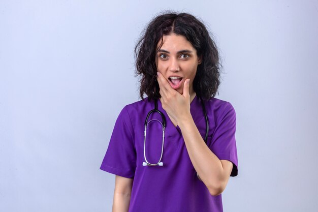 Enfermera joven en uniforme médico y con estetoscopio mirando sorprendido con la boca abierta con la mano en la barbilla