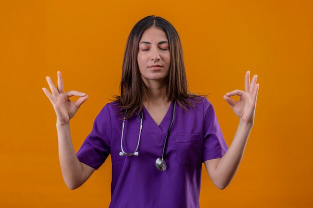 Enfermera joven en uniforme médico y con estetoscopio relajarse y sonreír con los ojos cerrados haciendo gesto de meditación con los dedos