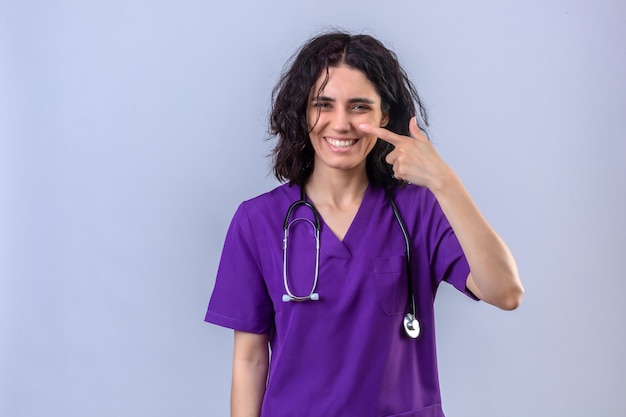 Enfermera joven en uniforme médico y con estetoscopio sonriendo con cara feliz apuntando con el dedo a la nariz de pie