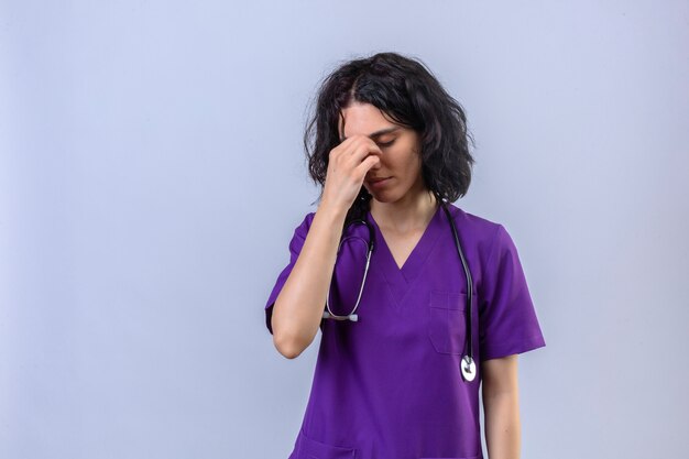 Enfermera joven en uniforme médico y con estetoscopio tocando la nariz entre los ojos cerrados destacó la sensación de fatiga de pie