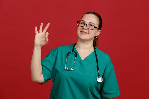 Enfermera joven con uniforme médico que usa anteojos con estetoscopio alrededor del cuello mirando a la cámara sonriendo feliz y positivamente haciendo el signo correcto sobre fondo rojo