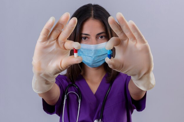 Enfermera joven vistiendo guantes de máscara protectora uniforme médico y con estetoscopio mostrando pastillas en las manos mirando confiado