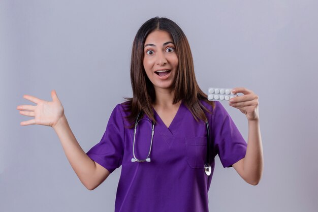 Enfermera joven vistiendo uniforme y con estetoscopio sosteniendo blister de píldoras presentando con el brazo de la mano hacia el lado salido y feliz