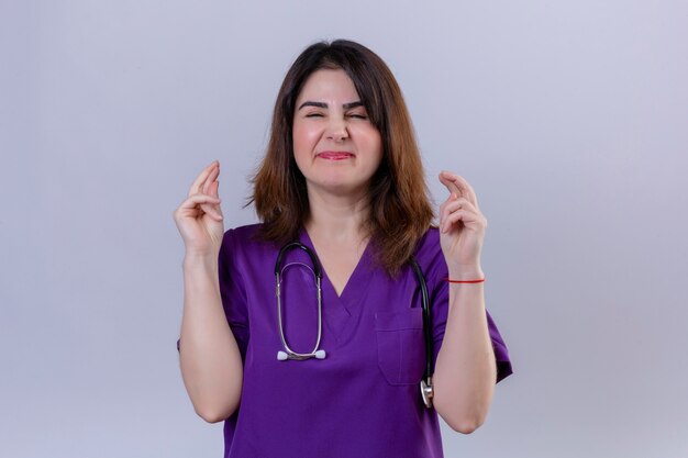 Enfermera de mediana edad con uniforme y con estetoscopio con los ojos cerrados levanta los dedos cruzados