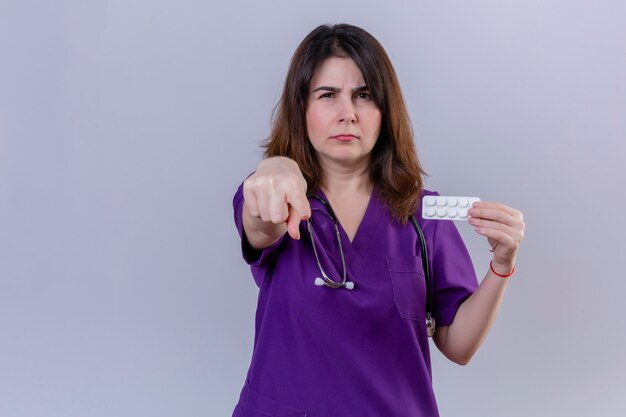 Enfermera de mediana edad con uniforme médico y con estetoscopio con ampolla con píldoras apuntando disgustado