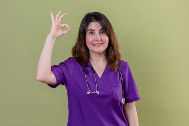 Enfermera de mediana edad con uniforme médico y con estetoscopio sonriendo alegremente haciendo bien firmar sobre pared verde aislado