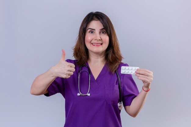 Enfermera de mediana edad vistiendo uniforme médico y con estetoscopio sosteniendo blister con pastillas mirando a la cámara con la cara hapy mostrando los pulgares para arriba sobre fondo blanco.