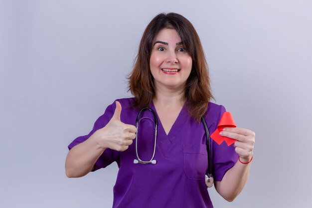 Enfermera de mediana edad vistiendo uniforme médico y con estetoscopio sosteniendo una cinta roja, un símbolo de la lucha contra el sida, mostrando los pulgares hacia arriba con la cara feliz de pie sobre fondo blanco.