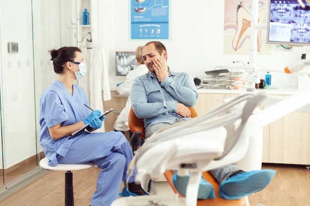 Enfermera médica examinando al paciente en paing escribiendo un problema dental en el portapapeles mientras está sentado en la oficina del hospital dental