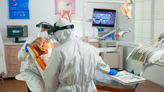 Enfermera y médico en traje de protección que trabajan en la unidad dental durante la pandemia de coronavirus que tratan a un paciente mayor. Asistente y médico de ortodoncia con mono, protector facial, mascarilla y guantes.