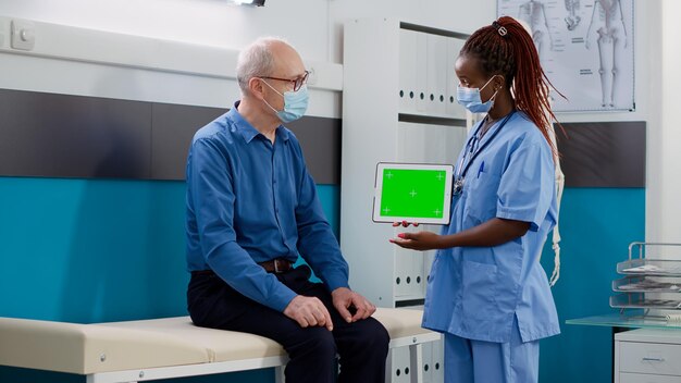 Enfermera mostrando tableta con pantalla verde a hombre mayor en gabinete médico. Mirando el fondo de cromakey aislado con plantilla de maqueta en blanco y espacio de copia durante la pandemia.
