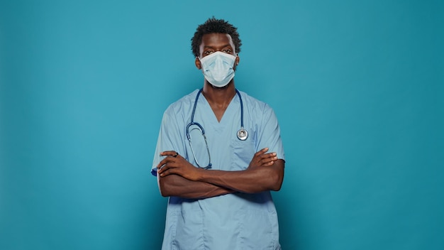 Enfermera de pie con los brazos cruzados mientras usa mascarilla para protegerse contra la pandemia del coronavirus. Asistente médico con uniforme sanitario y estetoscopio mirando a la cámara.