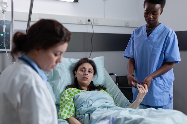 Enfermera que mide la saturación de spo2 usando un oxímetro de pulso en el dedo del paciente mientras el médico lee los resultados del laboratorio en una sala privada. Mujer atendida en cama de hospital con baja saturación de oxígeno mirando al médico.