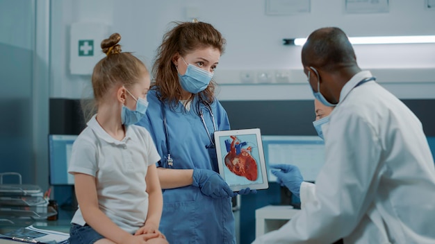 Enfermera que muestra una imagen de cardiología en una tableta digital, dejando que el médico explique el diagnóstico cardiovascular al niño y al padre. Asistente sosteniendo un dispositivo moderno con una imagen de la anatomía del corazón.