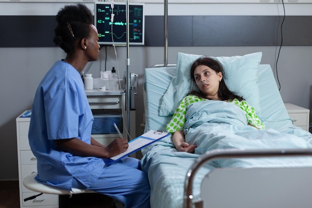 Enfermera sentada junto a la cama del paciente escribiendo en el portapapeles con historial médico durante la visita diaria consultando a una mujer con baja saturación de spo2. Persona en cama de hospital con signos vitales monitoreados durante el chequeo.