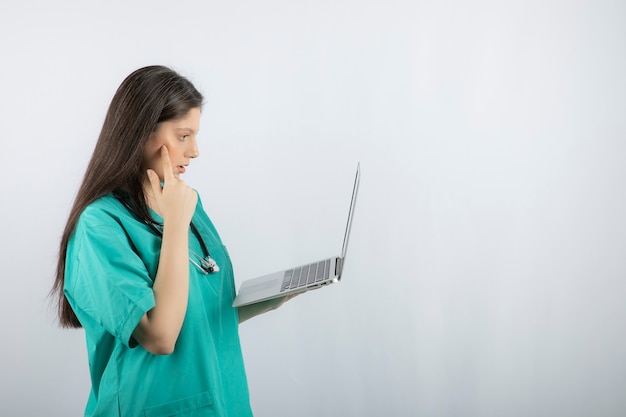 Enfermera de sexo femenino joven que mira algo en la computadora portátil sobre la pared blanca.