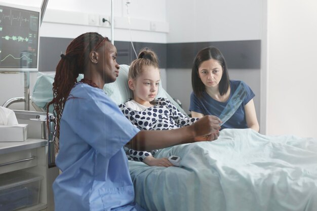 Enfermera sonriente pediátrica del hospital que muestra resultados de radiografía de niña enferma. Madre inquieta e hija sorprendida mirando la imagen de exploración de rayos X mientras está en la sala de recuperación del paciente.